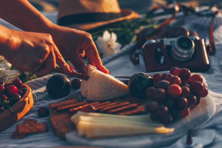 woman-slicing-cheese-wooden-cutting-board-with-cheese-fruits-it-camera-hat-flowers-beach (1)